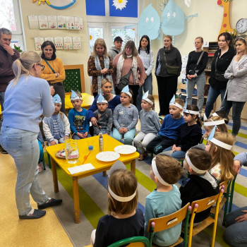 dzieci z w opaskach symbolizujących krople wody siedzą wokół stolika na którym pani prowadząca przeprowadza eksperymenty w tle widać grupę osób dorosłych 